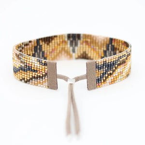 The JULIE ROFMAN - ORO BRACELET by JULIE ROFMAN JEWELRY is a woven piece adorned with a geometric pattern in shades of brown, beige, and black. It features handwoven Delica beads and includes a suede tie closure for adjustable sizing. The bracelet is displayed on a plain white background.