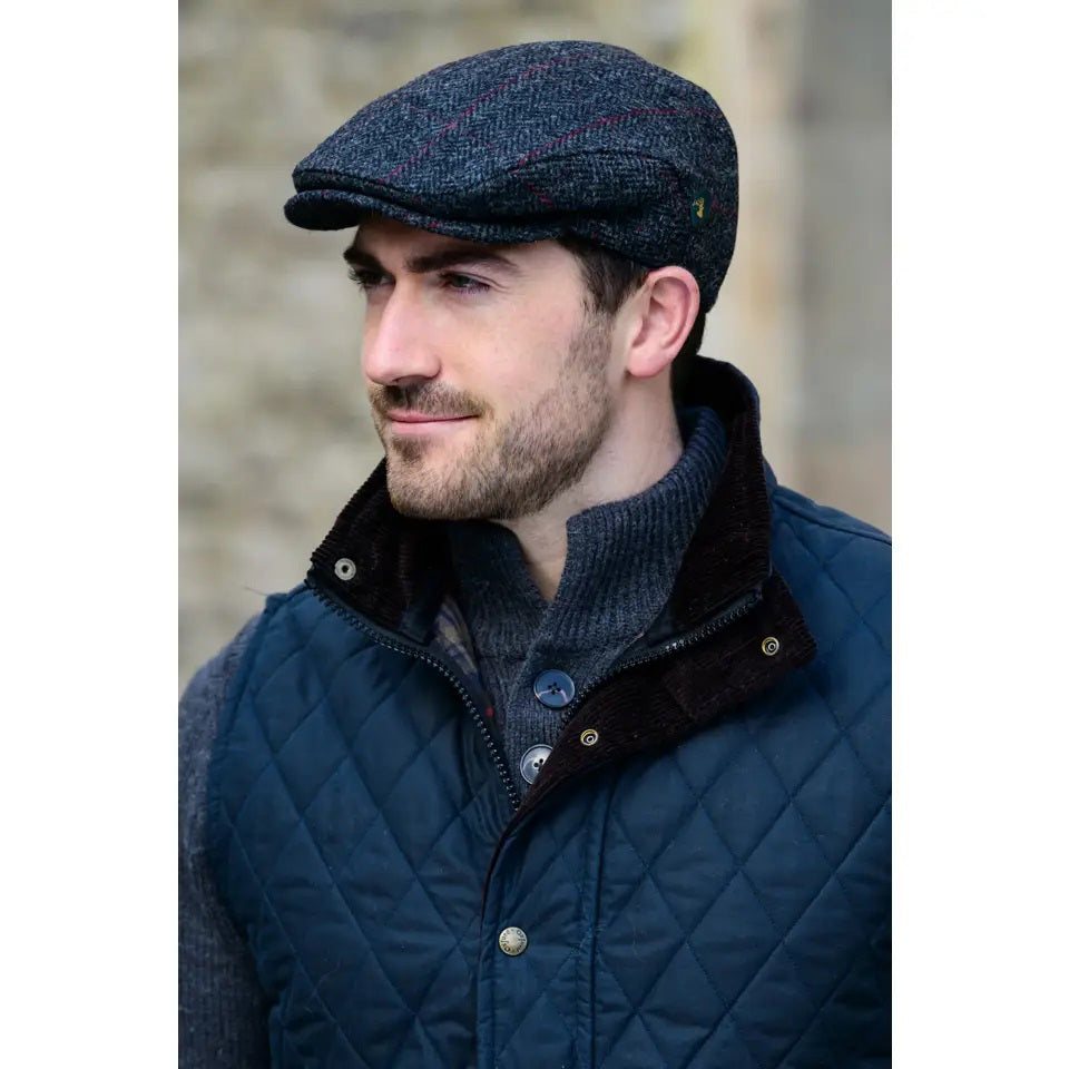 A man with a short beard, wearing the MUCROS WEAVERS - TWEED TRINITY CAP and a gray buttoned sweater with a navy quilted vest, stands outdoors against a blurred stone background.