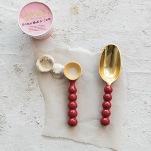 A container of Jeni's Gooey Butter Cake ice cream is opened on parchment paper. Two scoops, made with the Creative Co-op Enameled Aluminum Ice Cream Scoop that boasts a red & gold finish, sit beside two differently sized scoops. The surface is a light, textured material ideal for displaying this food-safe treat.