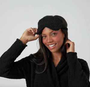 A person with long dark hair smiles while holding a black KASHWERE - EYE MASK above their eyes. They are dressed in a black, soft-textured robe and stand against a plain, light gray background, clearly prepared for restful sleep or travel relaxation.