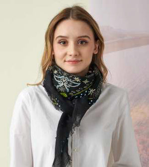 A young woman with light brown hair wears the STORIATIPIC Nelly Square Cashmere and Wool Embroidered Bandana Scarf, paired with a light gray top, standing before a softly colored nature-themed backdrop.