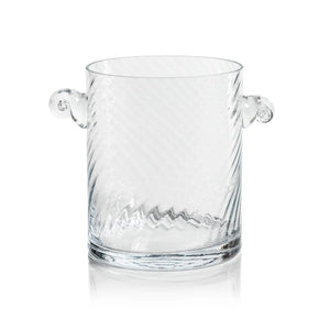 The BAGATELLE SWIRL ICE BUCKET by ZODAX is a clear glass, cylindrical ice bucket measuring 15.24 cm x 17.78 cm, adorned with subtle diagonal swirl patterns. It features two ornate looped handles on opposite sides and is showcased against a plain white background.