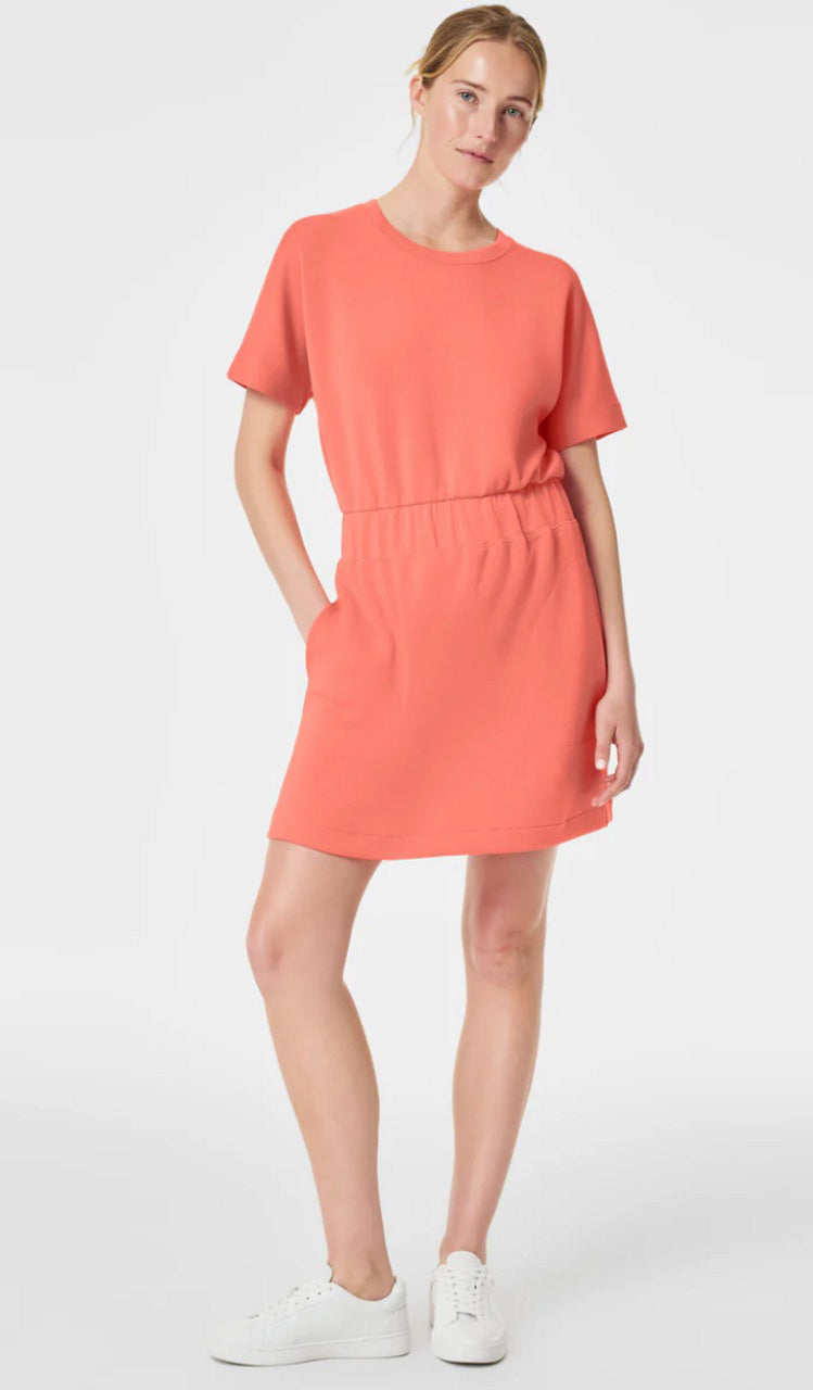 Wearing the SPANX Aire Essentials Cinched T-Shirt Dress in coral, a person strikes a pose with one hand in their pocket. The short-sleeve dress features a hidden elastic waistband and is paired with white sneakers, all set against a plain white background.