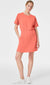 Wearing the SPANX Aire Essentials Cinched T-Shirt Dress in coral, a person strikes a pose with one hand in their pocket. The short-sleeve dress features a hidden elastic waistband and is paired with white sneakers, all set against a plain white background.