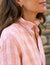 A close-up of a person wearing a FRANK & EILEEN - EILEEN Relaxed Button-Up Shirt in orange and cream plaid flannel linen. They have long, wavy brown hair and are turned slightly to the side, showcasing a delicate earring. The background is blurred and appears to be a natural outdoor setting.