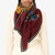 A person is wearing a cream-colored sweater with the INOUI EDITIONS - WESTERN SQUARE SCARF from INOUI, a large, burgundy wool scarf adorned with unique prints draped around their neck. The image focuses on the upper body, showcasing the intricate design of the scarf against a plain white background.