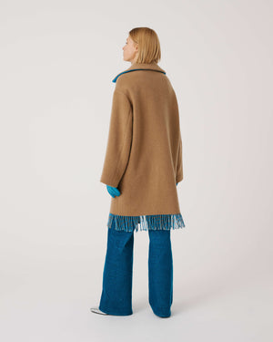 A person is wearing the SFIZIO - TWO TONED COAT WITH FRINGE by SFIZIO, featuring blue and brown fringe patch pockets and hem. They are also donning blue gloves and blue pants. The image is cropped to show from the shoulders down against a plain, off-white background.