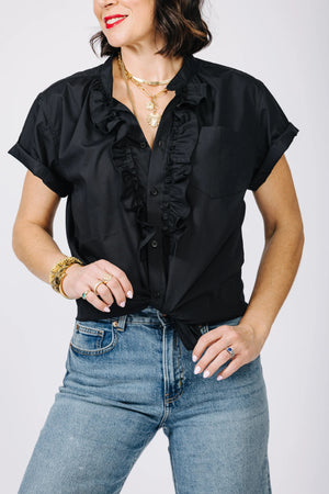 A person wearing the KMJ - KIKI SHIRT, a white ruffled blouse, and blue jeans stands with hands on hips, embodying a refined style. They accessorize with gold bracelets, rings, and a necklace. Their nails are neatly manicured and painted white. The background is plain.