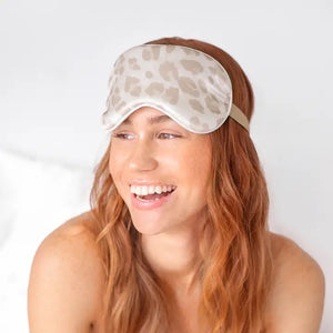 A LEOPARD SATIN SLEEP MASK from KITSCH, featuring a chic beige and white leopard print design, is displayed in front of its packaging. The packaging includes a cutout showcasing the sleep mask alongside an image of a woman wearing it. Text on the packaging reads: "The KITSCH Satin Eye Masks for deeper sleep," highlighting this luxurious satin product from a female-owned company.