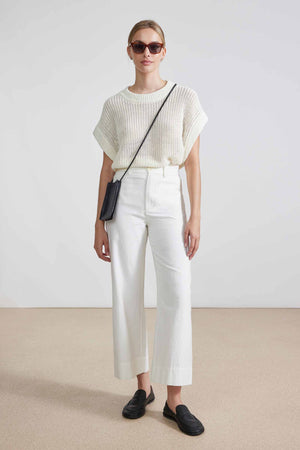 A person with hair in a bun is facing away, wearing sunglasses, a cream knit top, and the APIECE APART - CLASSIC MERIDA PANT that creates a sleek silhouette. Black shoes and a thin black crossbody strap complete the look against a light, minimalist setting.