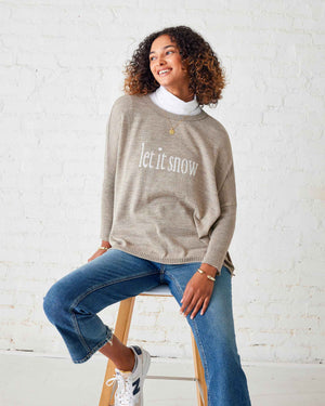 A person with curly hair is wearing the MERSEA - LET IT SNOW CATALINA CREWNECK SWEATER in beige over a white shirt and blue jeans, standing sideways against a white brick wall. They are smiling and looking at the camera.