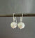 A pair of ANN LIGHTFOOT Freshwater Pearl Drop Earrings, showcasing round, lustrous grey freshwater pearls suspended from delicate sterling silver earwires, is pictured against a simple, dark background.