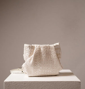 A black, textured handbag with a rectangular base and a gathered top is displayed on a white pedestal. The handbag, designed by DANIELA LEHAVI and named the TOKYO BALI BAG, features an alligator-like pattern and appears to be made of leather or a similar material. The background is plain and neutral-colored.