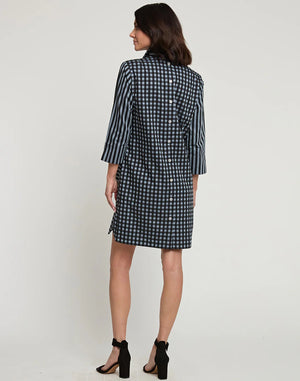 A woman stands posing sideways in a knee-length, loose-fitting HINSON WU AILEEN 3/4 Sleeve Dress featuring a classic black and tan stripe and gingham combo. She has shoulder-length dark hair and wears black high-heeled sandals. The background is a plain white wall.