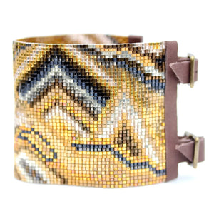 Introducing the JULIE ROFMAN - ORO CUFF by JULIE ROFMAN JEWELRY, a beaded bracelet that showcases an intricate geometric pattern in hues of gold, brown, blue, and white. The delica glass beads form a mosaic-like effect with abstract mountain and wave motifs in a stunning landscape design accented with 24kt gold-plated touches.