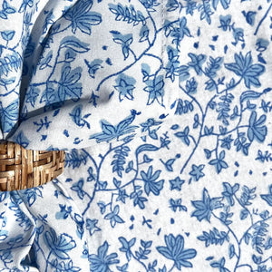 The CHRISTINA DICKSON HOME - WHIMSY FLORAL COTTON NAPKIN, an artisan-crafted blue and white floral fabric made in India, is tied in a knot over a circular rattan mat. Its delicate leaf and flower design complements the mat's textured pattern.