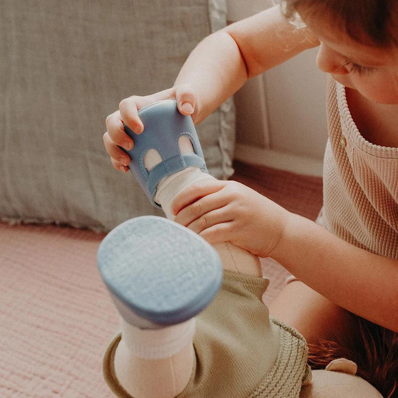A close-up of a baby wearing OLLI ELLA USA DINKUM DOLL SHOES in Mallow Pink and white socks. The baby is seated on a soft, pale pink textured blanket, dressed in light brown clothing. Only the lower half of the baby&#39;s body is visible.