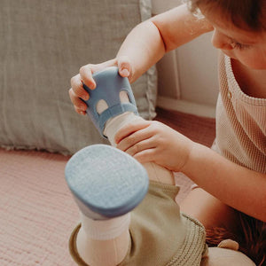 A close-up of a baby wearing OLLI ELLA USA DINKUM DOLL SHOES in Mallow Pink and white socks. The baby is seated on a soft, pale pink textured blanket, dressed in light brown clothing. Only the lower half of the baby's body is visible.