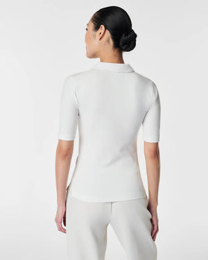 A woman in the SPANX - STRETCH RIB POLO TOP, a brown, ribbed, short-sleeve shirt with a button-down front, stands with her arms relaxed. The plain white background accentuates the top's stretchy ribbed fabric and body-hugging fit.