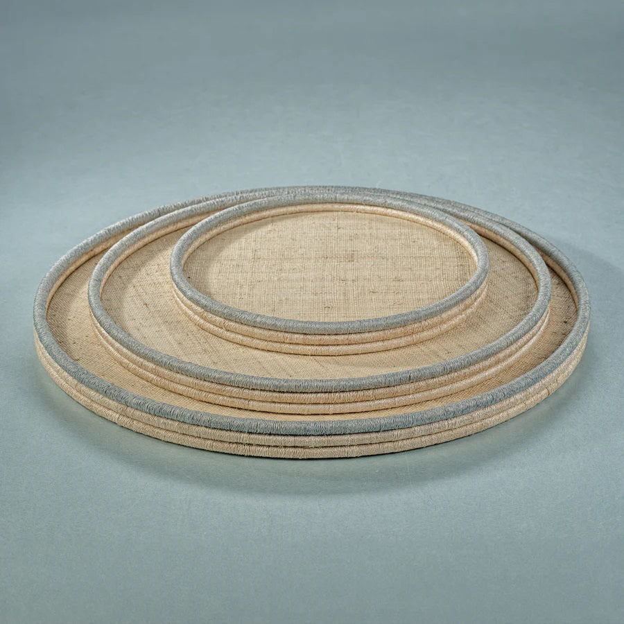 A round, flat **ZODAX** tray with a woven texture sits against a plain white background. The **COILED RATTAN AND ABACA ROUND TRAY**, measuring 24.25 in x 1.5 in, features a beige surface with slightly raised edges encased by a thin, blue border. The design is simple and rustic.