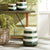 The NAPA HOME AND GARDEN - LIA STOOL is a whimsical ceramic with a tapered cylindrical shape featuring green and white horizontal stripes in gradient patterns. Varying shades of green mimic natural elements, creating enchanting hue variations.