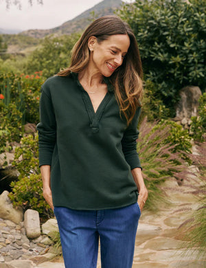 A person with long brown hair wearing a dark green FRANK & EILEEN - Patrick Popover Henley in Triple Fleece Evergreen stands outdoors. They are adjusting the collar of their top with both hands. The background is lush with greenery and soft-focus plants.
