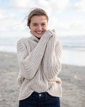 A woman with curly hair stands against a light-colored wall, wearing the MERSEA - BEACHSIDE TURTLENECK from MER SEA over a white shirt, black pants, and black flat shoes. Beside her is a woven cylindrical ottoman. She gazes thoughtfully to the side with a relaxed posture.