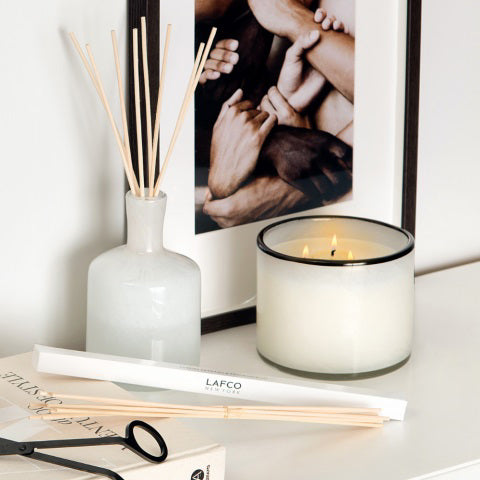 A LAFCO Champagne Penthouse 3 Wick Candle with a black rim burns next to a white vase holding essential oil-infused reed diffusers. A photo of intertwined hands rests in the background, while a box labeled "LAFCO" and wick trimmers lie nearby.