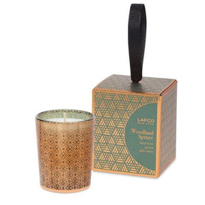 A LAFCO - WOODLAND SPRUCE VOTIVE candle in a patterned glass holder is placed beside its matching box. The box, adorned with green and gold geometric designs and a black ribbon handle, is labeled "LAFCO New York Woodland Spruce." The partially transparent glass of the candle reveals the wax inside, evoking the scent of an evergreen spruce.