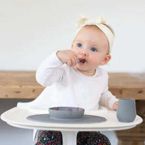 A toddler wearing a white headband and white shirt is sitting in a high chair, self-feeding with an infant spoon. There are a small bowl and an EZPZ Tiny Spoon 2pk - Gray on the tray in front of the child. The background features a wooden table and a white wall.