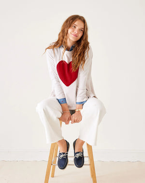 A smiling person with long hair wears the MERSEA Catalina Long Sleeve Slub Tee with Heart, over a blue button-up shirt. Paired with white pants, they stand against a plain background, capturing casual elegance effortlessly.