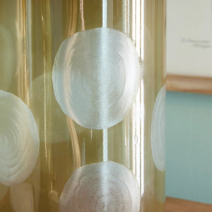 A table in a room with a light blue wall features whimsical lighting, including the NORMAN LAMP by NAPA HOME AND GARDEN, which has a taupe glass base and a white linen shade. The table is adorned with two brass bird statues and a small stack of books. Framed botanical prints hang on the wall.