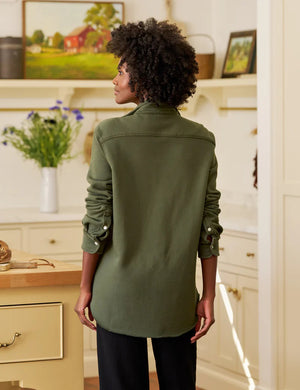 In a sunny, rustic kitchen, a woman smiles while wearing the FRANK & EILEEN EILEEN Relaxed Button Up in Triple Fleece Caper, paired with black wide-leg pants and sandals. She touches her hair as sunlight fills the cozy space.