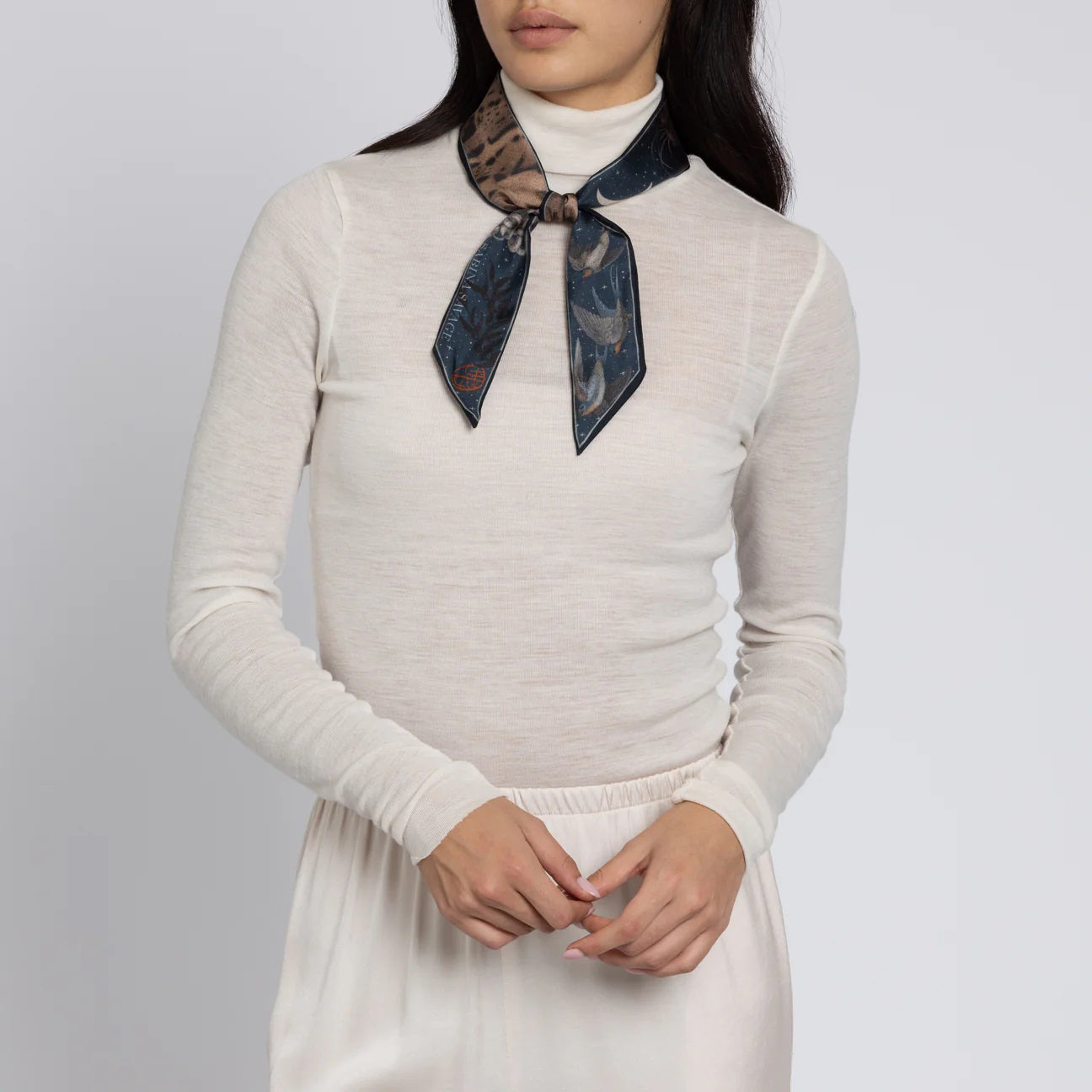 A person wearing a white turtleneck and a dark patterned neck scarf, reminiscent of the elegance found in the SABINA SAVAGE - THE LUNAR LEOPARD SILK RIBBON by SABINA SAVAGE ENGLAND, stands against a neutral background. Their hands are gently clasped in front.