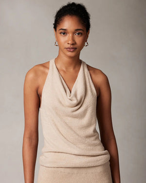 A woman with curly hair tied in a low bun is wearing the KASHWERE COWL NECK TOP by KASHWERE, paired with a matching knit skirt. She stands against a neutral background, facing to the left and showcasing the back of her outfit, which hints at athleisure vibes.