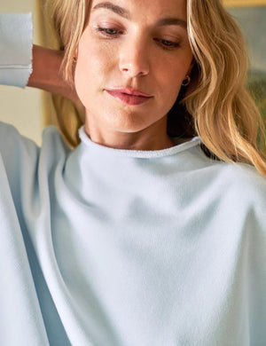 A woman with long hair, wearing a FRANK & EILEEN Effie Long Sleeve Funnel Neck Capelet in Ice and jeans, smiles over her shoulder. She stands in a room featuring a wooden sideboard, books, a candle, and a framed painting on the wall.