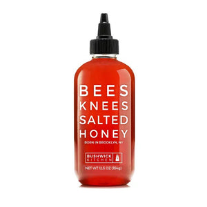 A red bottle of BUSHWICK KITCHEN - 'BEES KNEES' SALTED HONEY is shown. The label reads "BORN IN BROOKLYN, NY" and "NET WT 12.5 OZ (354g)." This gluten-free treat features wildflower honeys perfected with a hint of salt and comes with a black pointed cap.