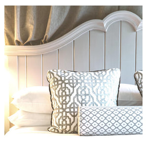 A close-up of a neatly made bed featuring the FEARRINGTON LIFESTYLE BEDDING COLLECTION - KING BEDDING EXPERIENCE from FEARRINGTON HOUSE. The white linens are complemented by two patterned pillows: the larger pillow boasts a geometric design in grey and white, while the smaller cylindrical bolster pillow features a diamond pattern in grey and white. A light-colored wooden headboard completes this luxurious sleep experience.