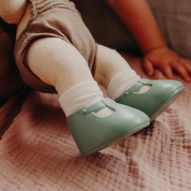 A close-up of a baby wearing OLLI ELLA USA DINKUM DOLL SHOES in Mallow Pink and white socks. The baby is seated on a soft, pale pink textured blanket, dressed in light brown clothing. Only the lower half of the baby&#39;s body is visible.