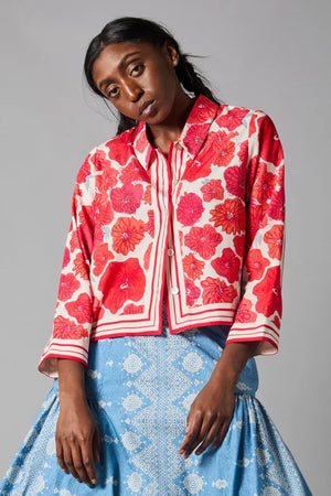 A person with dark braided hair wears the HOPE FOR FLOWERS - SCARF PRINT SHIRT, showcasing red and white floral patterns with striped accents. This loose-fitting blouse, crafted from sustainable REFIBRA fabric, is paired with a blue skirt featuring lace details against a plain gray background.