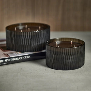 A cylindrical, dark green candle with a textured surface sits next to a matching dark brown, wood-textured box with a gold label. The box lid reads "ZODAX." The candle features five wicks and boasts fragrance notes of Cedar Leaf and Hinoki Cypress, with the box label reading "5 WICK CANDLE - CEDAR LEAF & HINOKI CYPRESS" to indicate the scent.