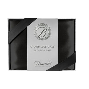 A black silk pillowcase is neatly packaged in a sleek black box with a white label that features an elegant "B" logo at the top. The label reads "BRANCHE - CHARMEUSE PILLOW CASE SILK: Sleep in Luxury. Wake up Beautiful," while promoting skin hydration and enhancing hair care.