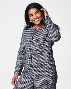 A person with long hair sits casually on a light-colored chair wearing the SPANX PONTE JACQUARD JACKET, which is a black and white checkered suit adorned with gold-finished buttons. They are gazing to the side, with one arm resting on the chair's armrest and the other in their lap.