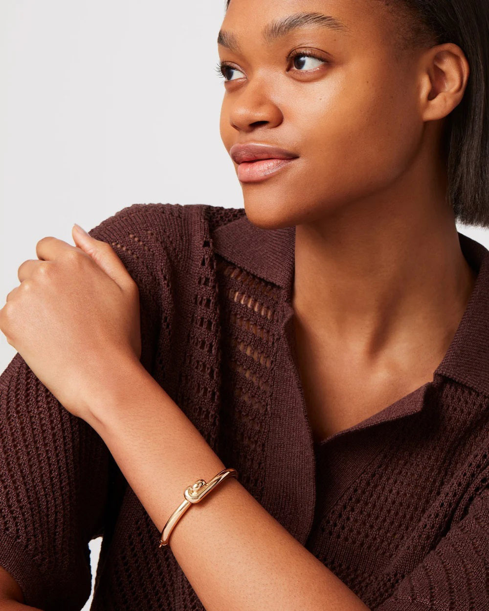 A woman in a brown knit sweater wears a gold Maeve Bangle by JENNYBIRD, featuring circular details, as she looks sideways. Her left hand rests on her opposite shoulder against a plain light gray background.