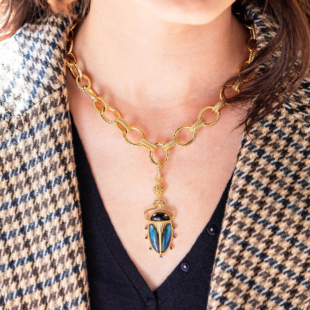 The CAPUCINE DE WULF - SCARAB PENDANT BLUE LABRADORITE/BLACK AGATE by CAPUCINE DE WULF features a gold scarab pendant with a central black agate stone, flanked by iridescent blue wings. The elaborate design includes a detailed floral top and small blue labradorite gemstones encircling the lower edge.