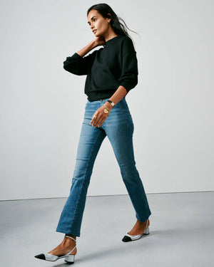 A person stands against a plain backdrop, confidently showcasing their style in SPANX's KICK FLARE JEAN, made with 4-way stretch denim. They pair it with a black sweater and black-and-white heels, their long dark hair cascading as they pose with one hand in their pocket.