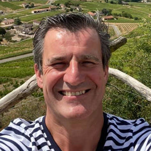 A person smiles in a selfie, donning a striped shirt against lush vineyards and village. The sunlit landscape showcases a truly Authentique Vin Wine Dinner experience with FEARRINGTON VILLAGE's French wines.