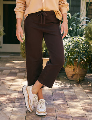 A person with long hair stands outdoors on a stone path, dressed in a dark outfit with FRANK & EILEEN's Catherine Favorite Sweatpants in Irish Chocolate and light brown shoes. Surrounded by lush greenery and with a modern building in the background, sunlight filters through to create a warm atmosphere.