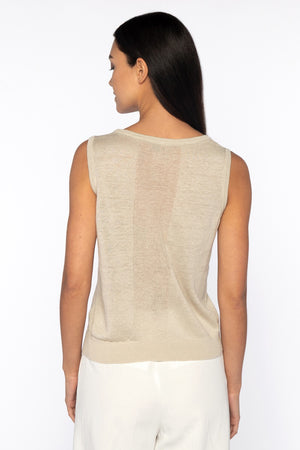 A woman with long dark hair stands against a white background, embodying elegance in a versatile KINROSS ensemble. Her serene and neutral expression complements the timeless essential look created by the KINROSS CASHMERE sleeveless tank in linen and white pants, hands rested casually in pockets.