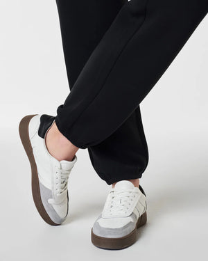 Dressed in SPANX - ARESSENTIALS JOGGER pants crafted from lightweight fabric, a person stands with hands in pockets, pairing them with a matching hoodie. They complete their look with white sneakers featuring brown soles. The plain white background accentuates their outfit's style and comfort.